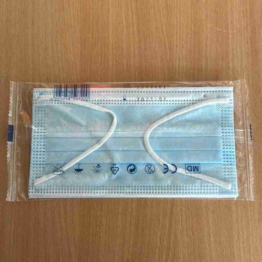 Einweg-Gesichtsmaske in transparenter Plastikverpackung auf Holzoberfläche, mit sichtbaren Symbolen für medizinische Verwendung, CE-Kennzeichnung und Haltbarkeitsdatum.